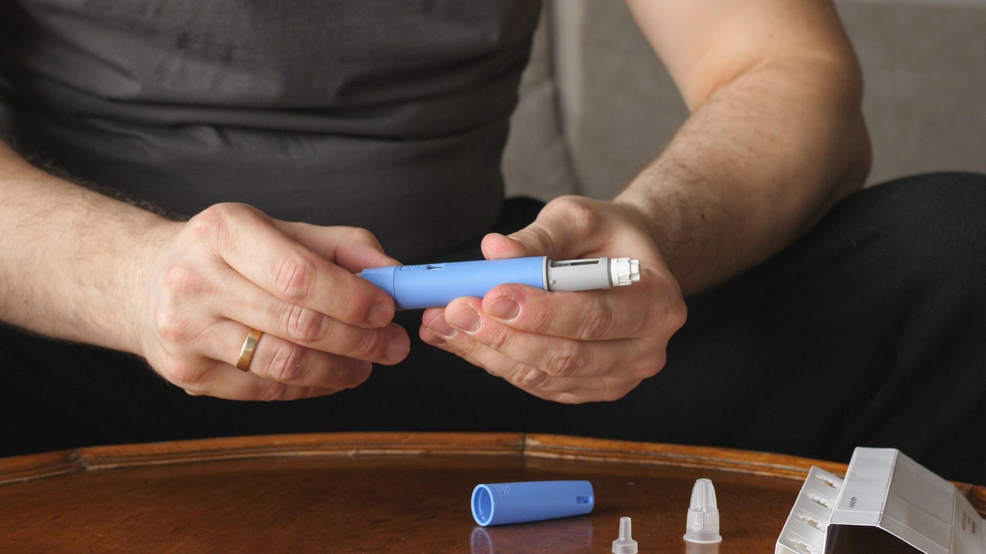 A person holding a blue pen injector, preparing it for use, with additional pen components on a wooden table.