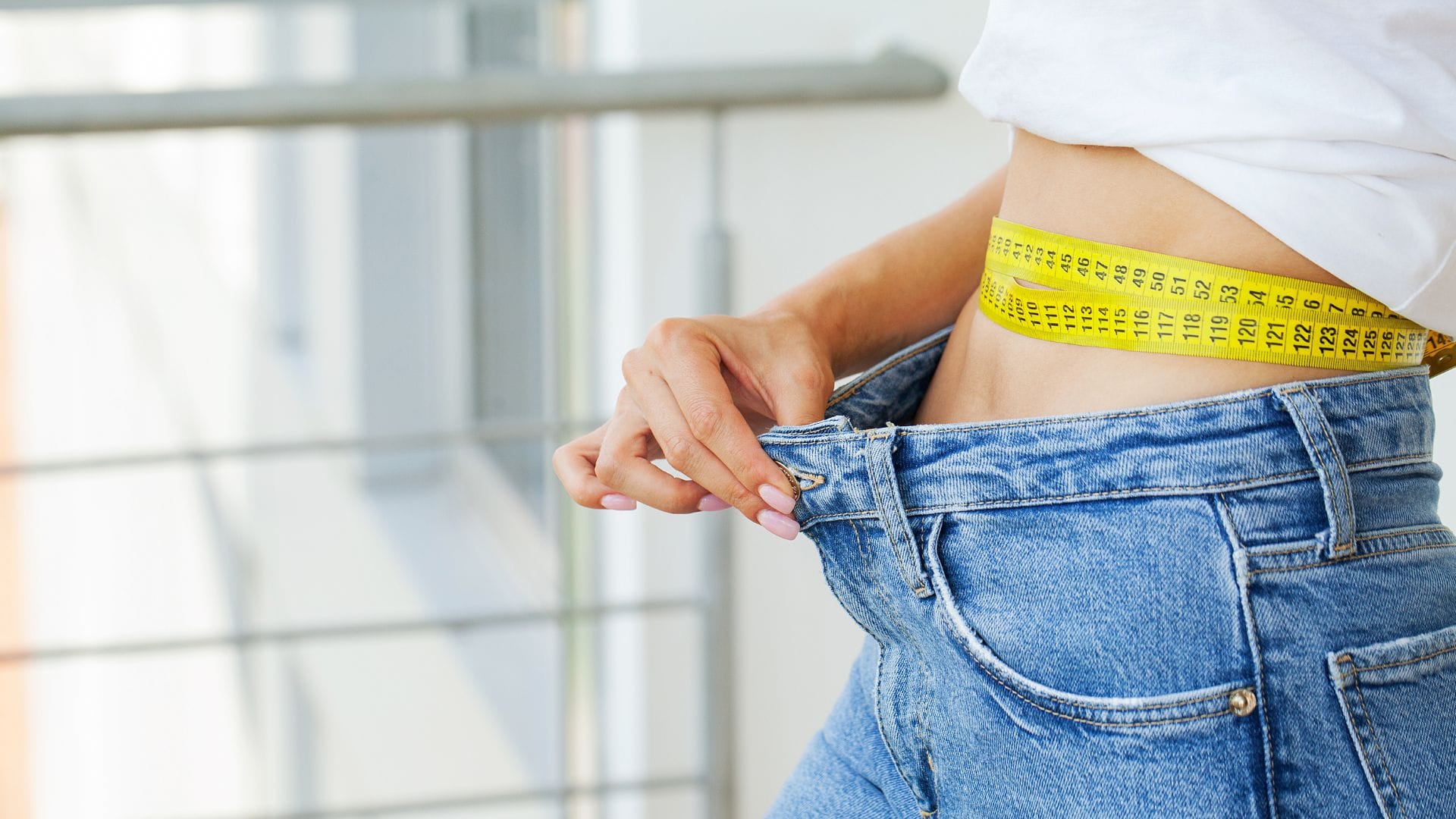 A woman measuring her waist with a yellow tape measure while wearing loose-fitting jeans, indicating weight loss.