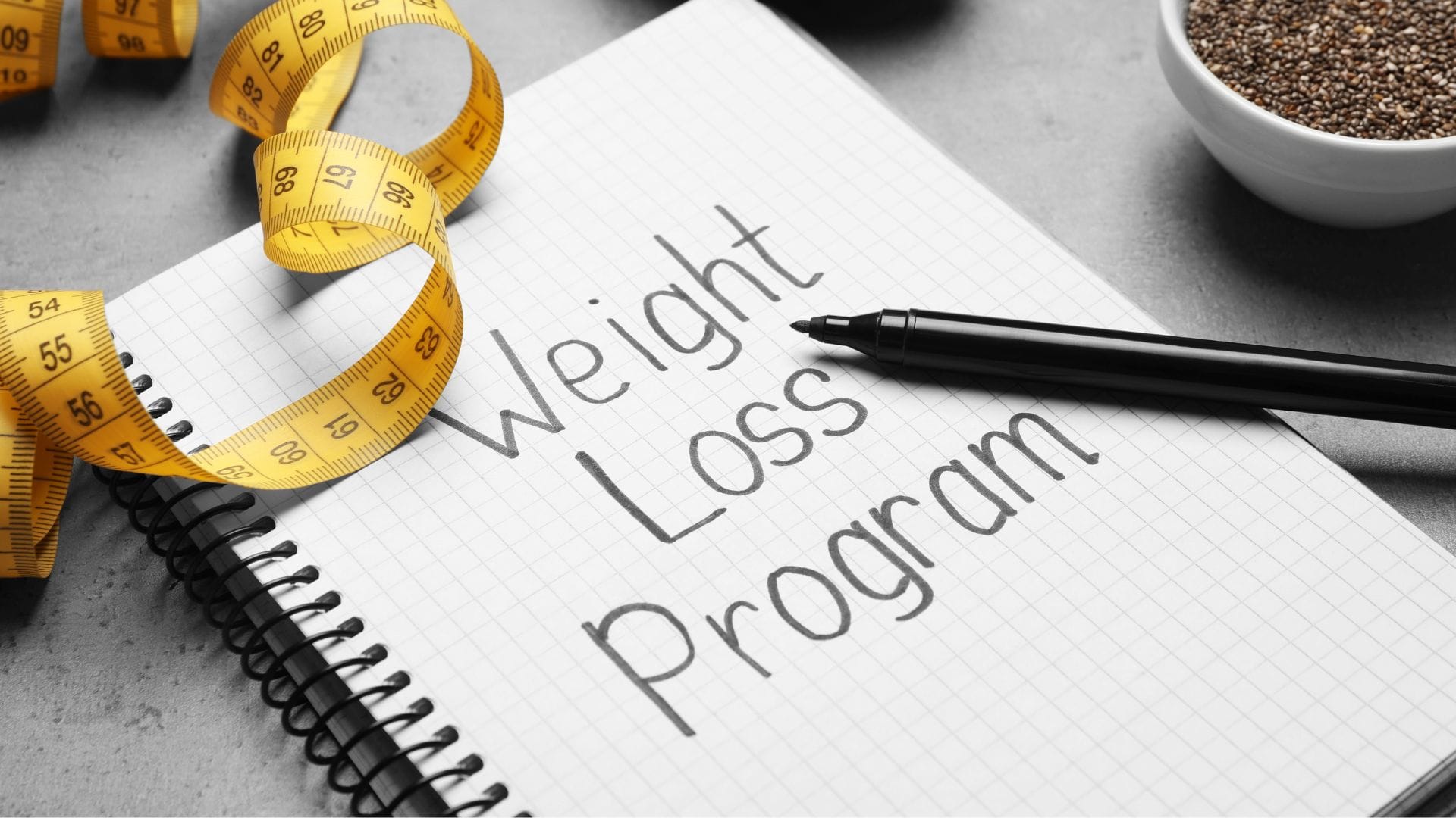  A spiral notebook with "Weight Loss Program" written on it, accompanied by a black pen and a yellow measuring tape.