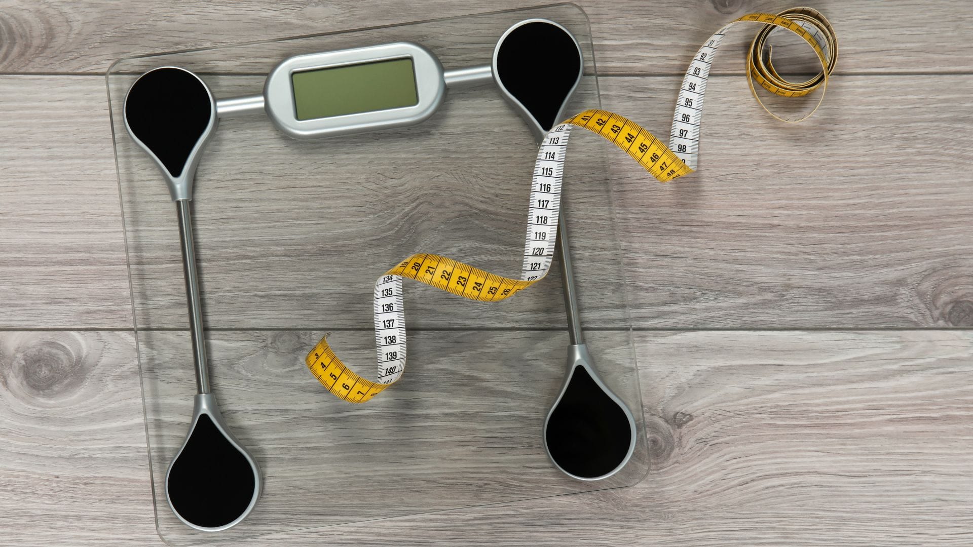 A digital weighing scale with a coiled yellow measuring tape on a wooden floor.