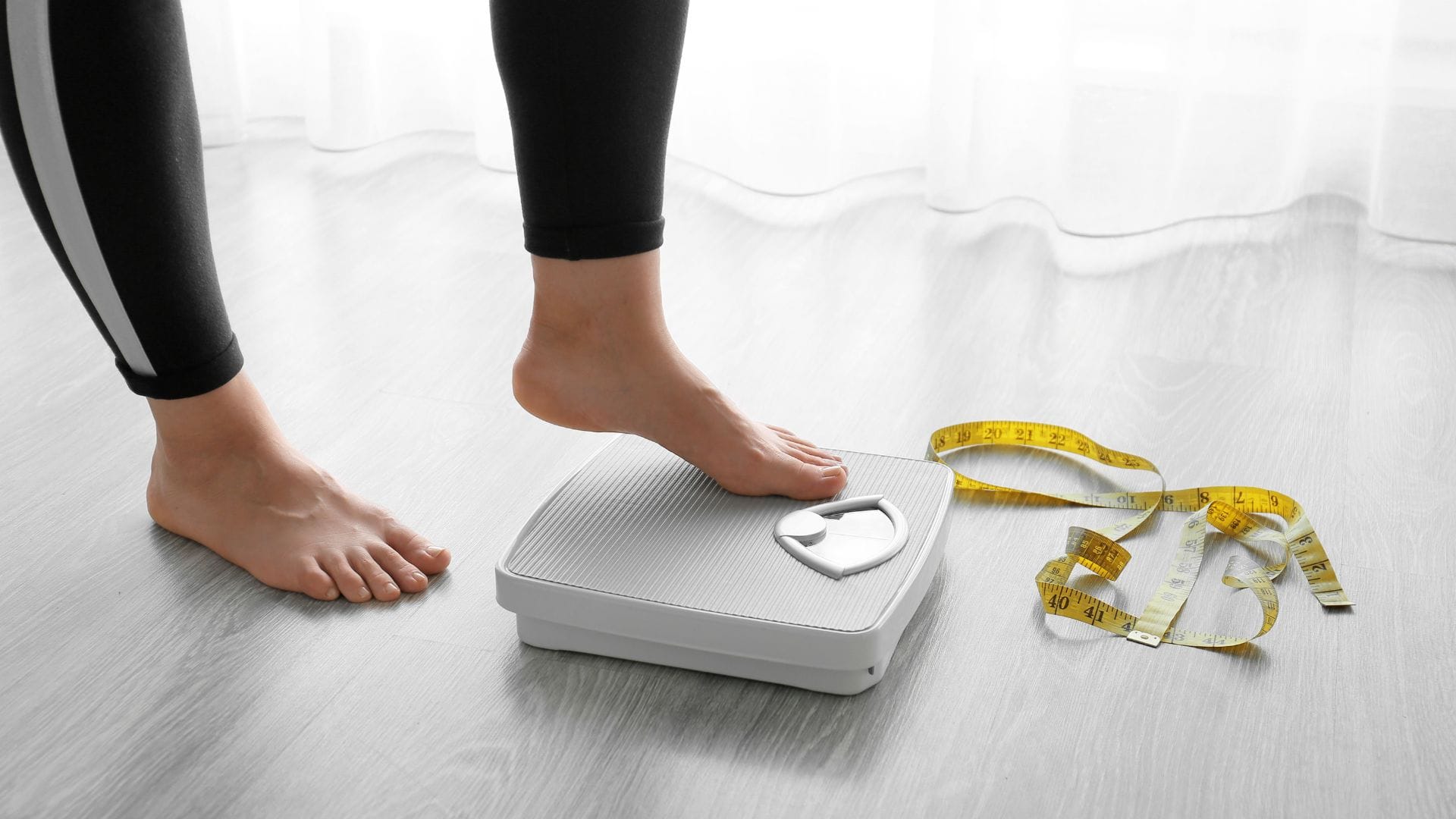 A person stepping onto a weighing scale with a tape measure on the floor.