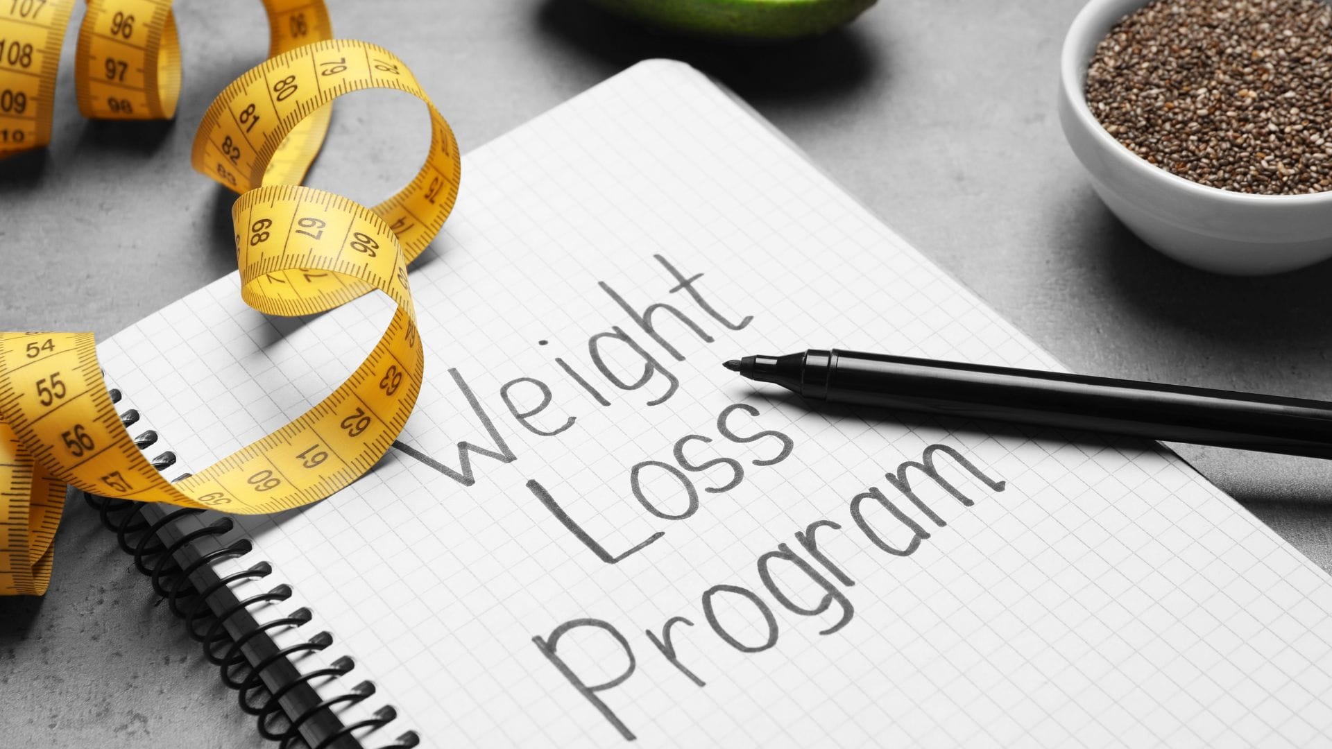 A notebook with "Weight Loss Program" written on it, alongside a yellow measuring tape and a pen.