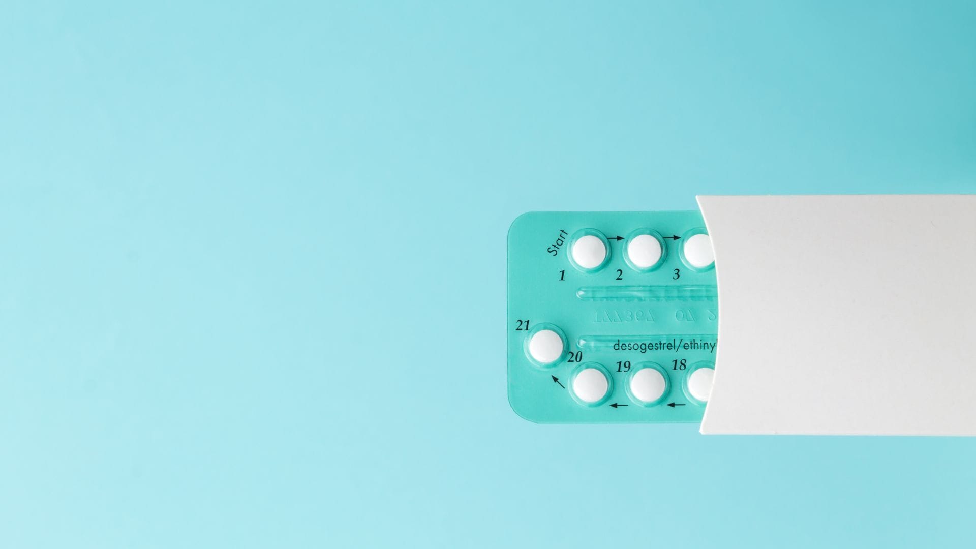 Blister pack of desogestrel and ethinyl estradiol birth control pills partially pulled out of white paper sleeve, set on a light blue background.