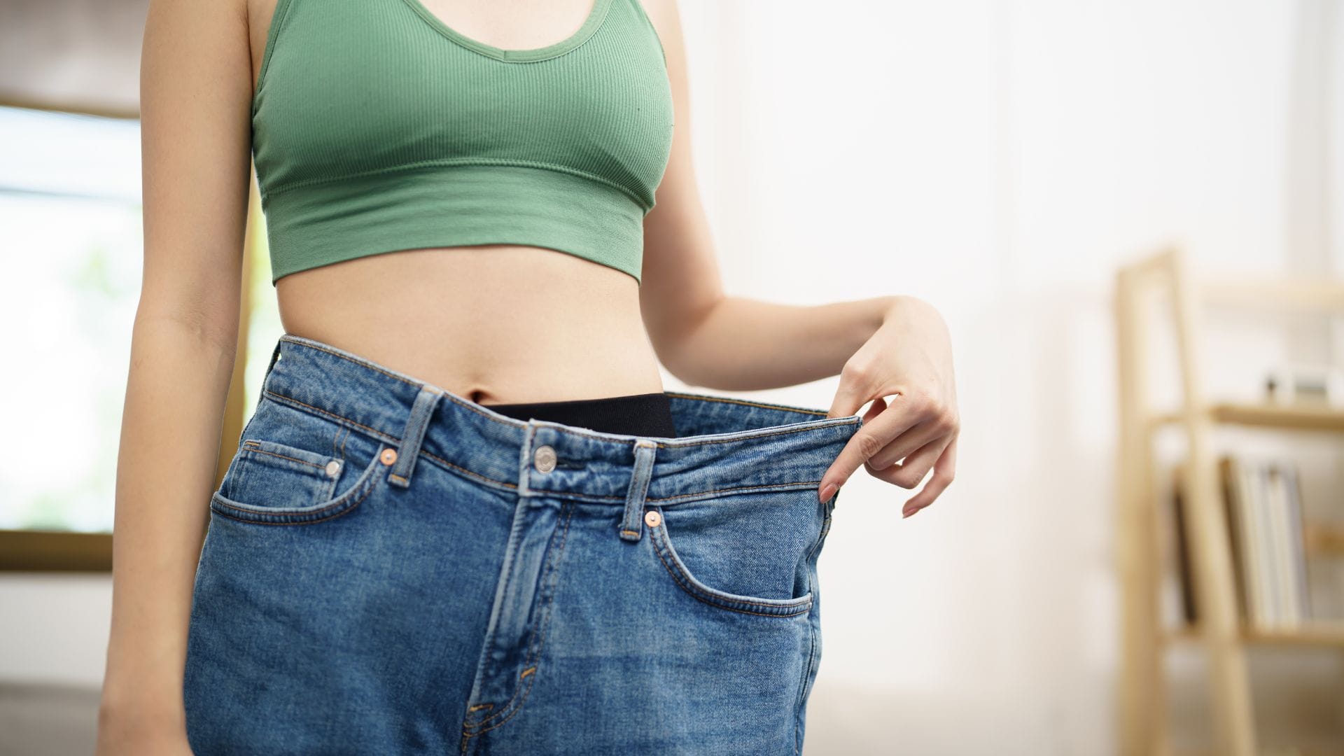 A person in a green crop top holding out loose-fitting jeans, illustrating weight loss.