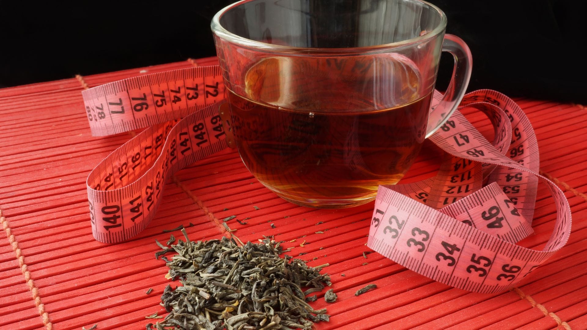 A cup of tea with a pink measuring tape and dried tea leaves on a red surface.
