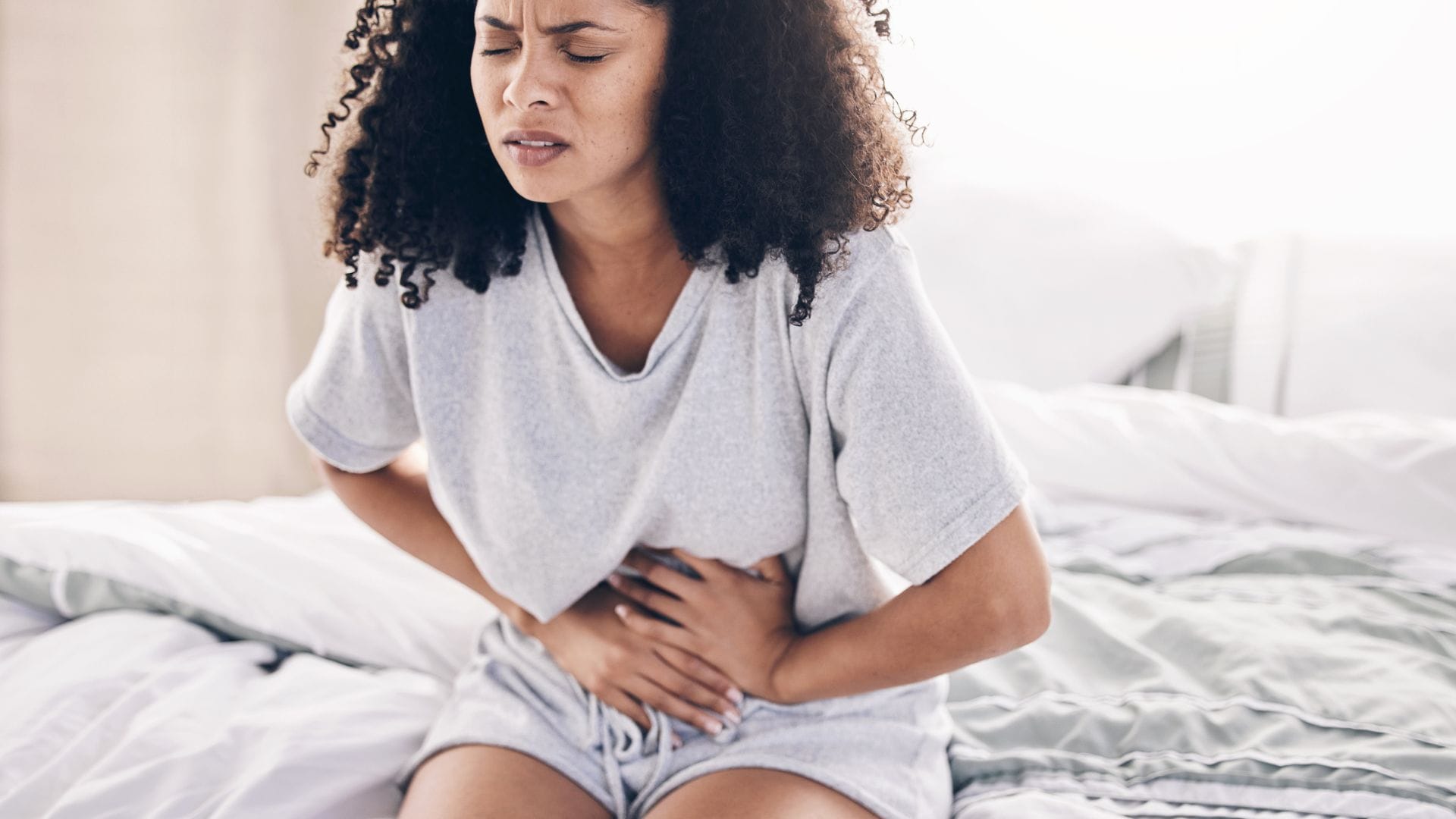 Woman sitting on a bed, clutching her abdomen in pain.