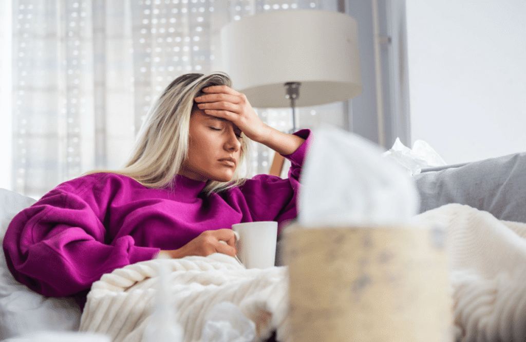 Woman resting on a sofa with her hand on her forehead, holding a mug while experiencing headache or migraine symptoms.
