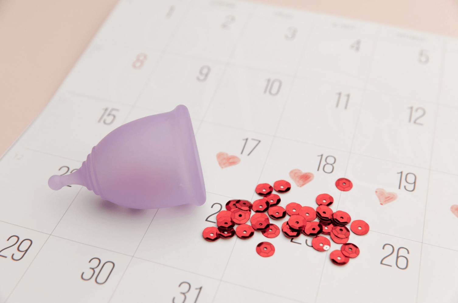 Purple menstrual cup placed on a monthly calendar beside red confetti and heart illustrations, representing period planning and menstrual cycle management.