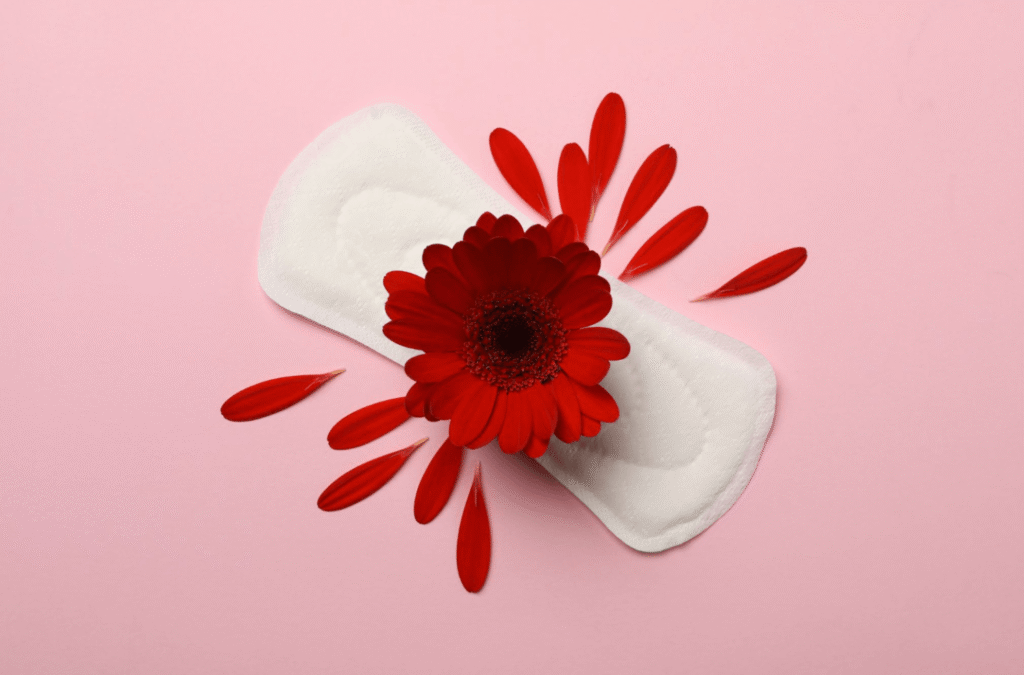 White sanitary pad with a red flower and scattered petals on a pink background, symbolising menstrual care and feminine hygiene.