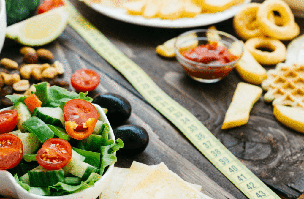 Bowl of fresh salad beside high-calorie foods and a measuring tape, illustrating the challenge of maintaining extremely low-calorie diets.