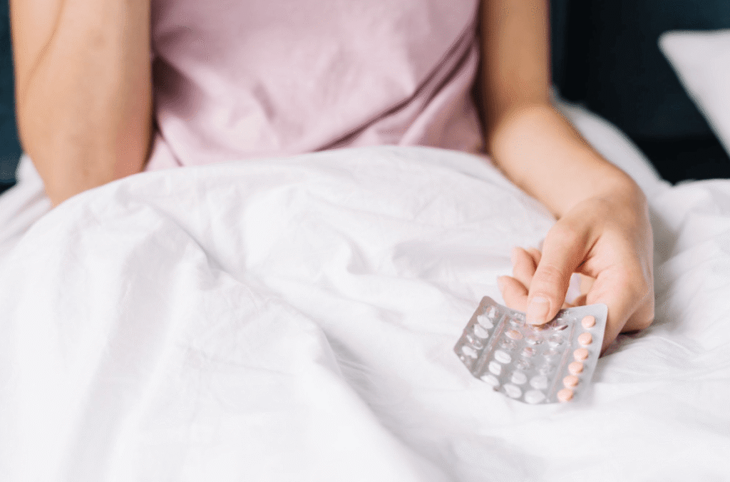 Person sitting in bed holding a blister pack of the morning after pill on white bedding.