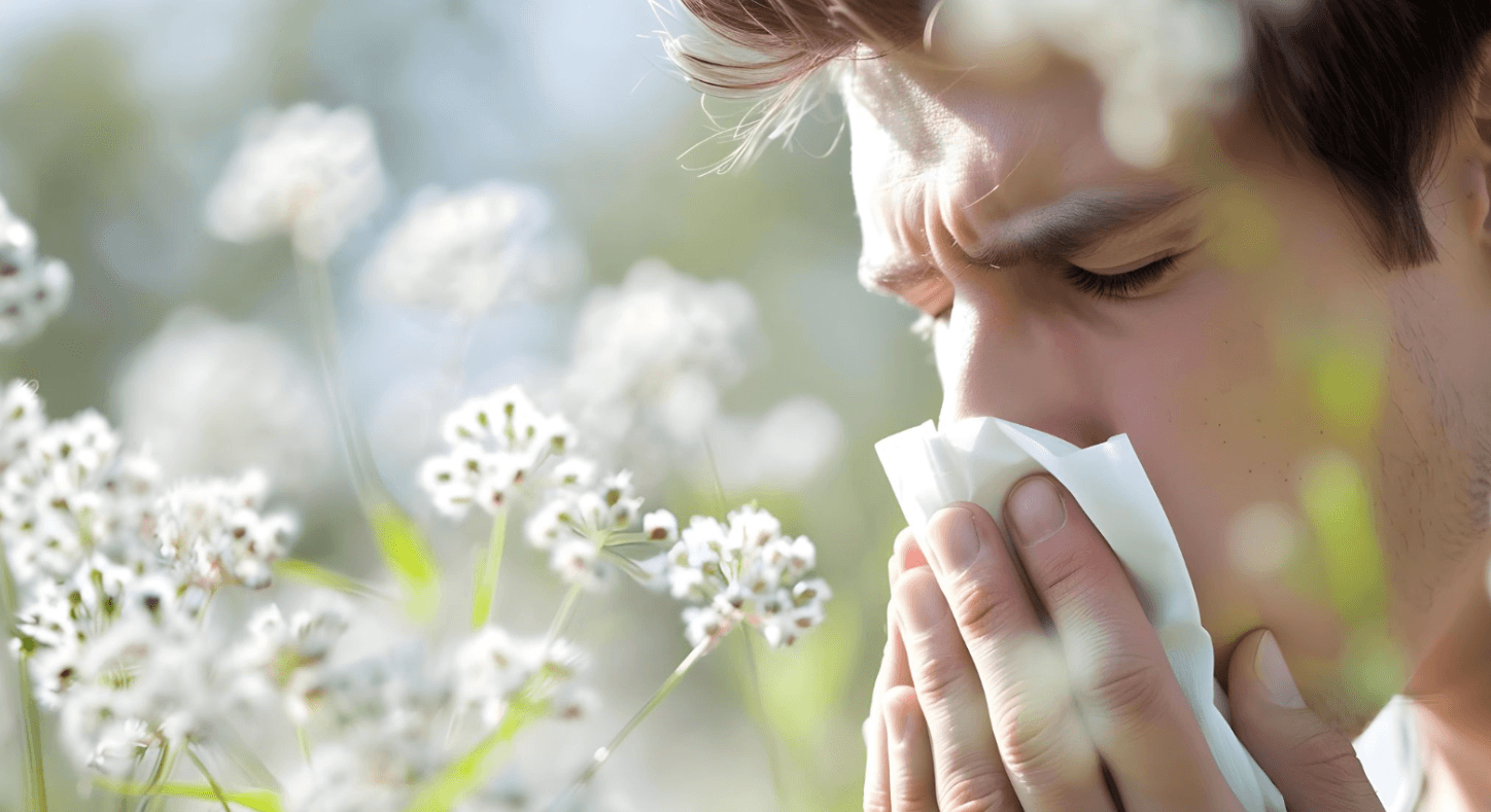 Man sneezing into a tissue outdoors among flowering plants during hay fever season.
