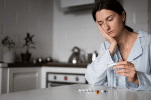 Woman sitting at a kitchen table with a glass of water and migraine tablets, feeling unwell before taking medication.