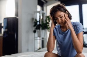 Person sitting with hands on their temples, experiencing migraine pain and discomfort in a bright indoor setting.