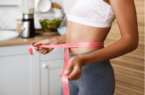 Woman measuring her waist with a tape measure while following a weight loss journey at home.