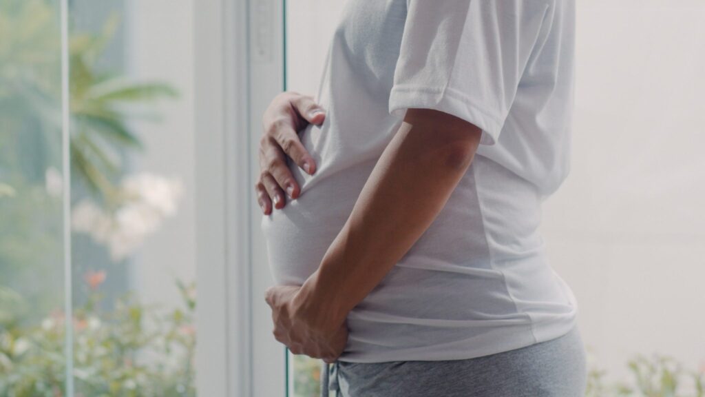 Pregnant woman gently holding her belly while standing beside a bright window.