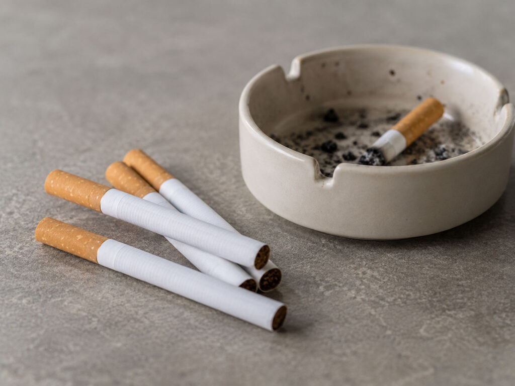 Several cigarettes lying beside a ceramic ashtray containing cigarette ash and a discarded cigarette.